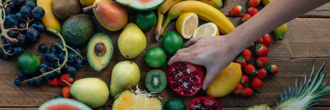Colorful variety of fresh fruits arranged on a wooden surface with a hand selecting a pomegranate.
