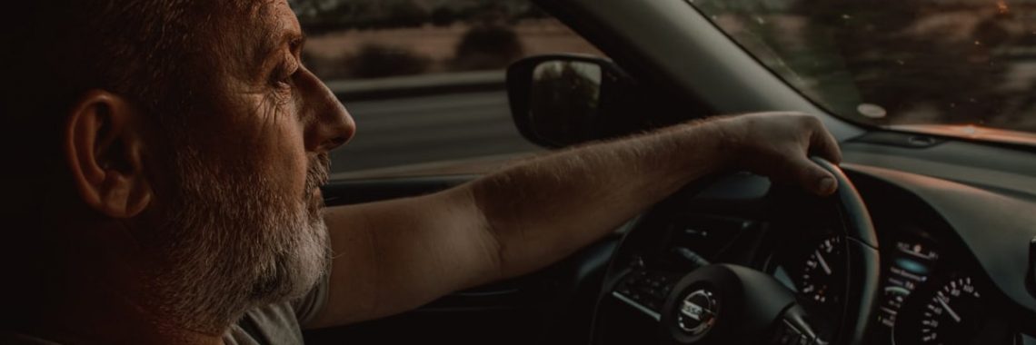 Elderly man driving a Nissan car during sunset, depicting travel and contemplation.